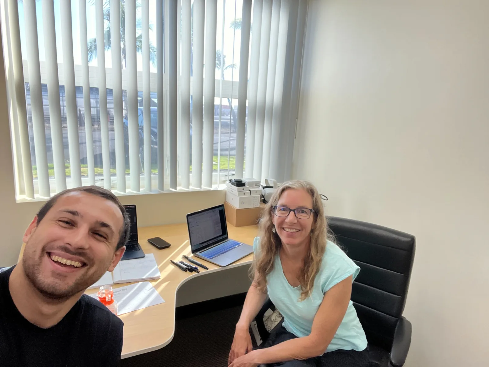 Ryan Westerberg and Tina Flower at a desk in their Kona office, laptops open, palms visible through the window behind them.